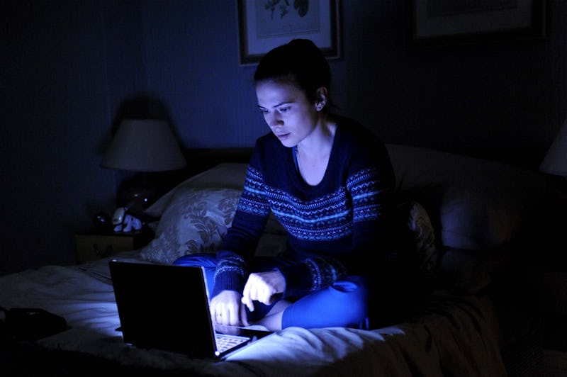 Young woman looking at laptop screen in a dark room while sitting on the bed 