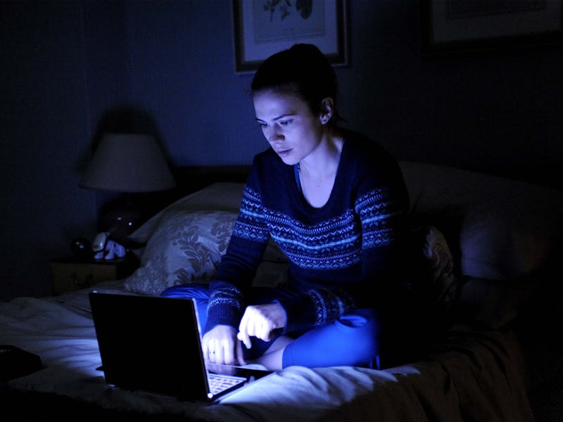 Young woman looking at laptop screen in a dark room while sitting on the bed 