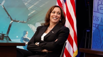 Kamala Harris smiling in a black blazer and white shirt with an American flag behind her