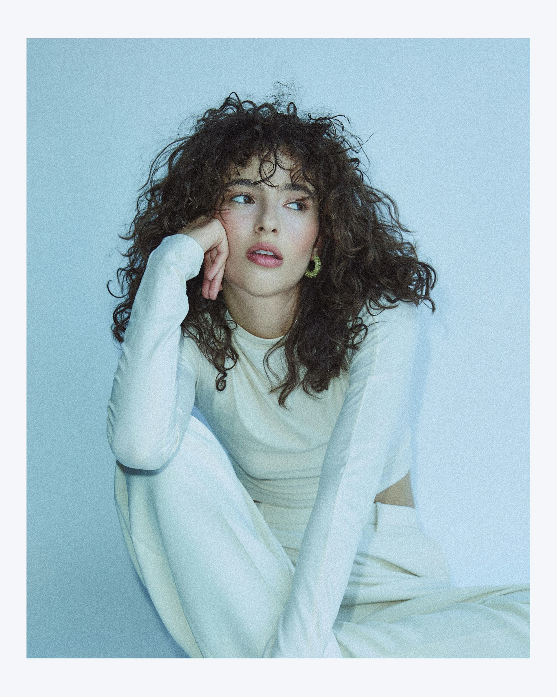 Nina Bloomgarden wearing an all white outfit, curly hair and intense blush along with hoop earrings 