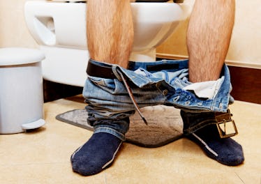 The lower portion of the legs with pants at ankles as a man sits down on a toilet to pee.