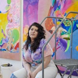 The artist Ilana sitting on a ladder in her studio, in front of one of her very colorful works.