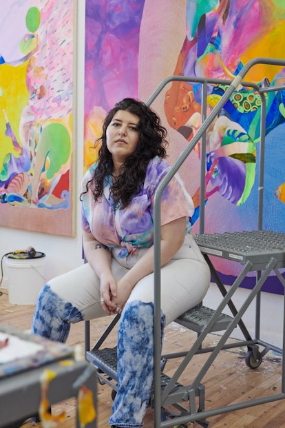 The artist Ilana sitting on a ladder in her studio, in front of one of her very colorful works.