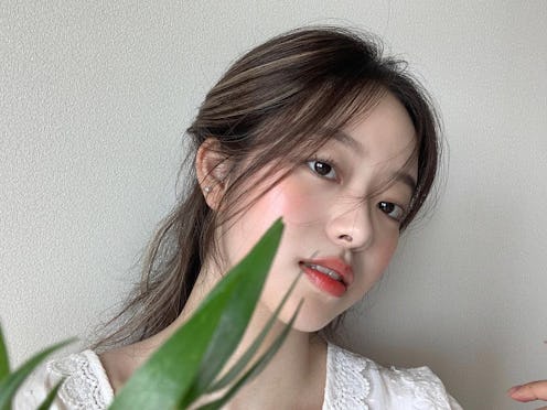Woman with glossy peachy makeup posing against a white wall with a leaf
