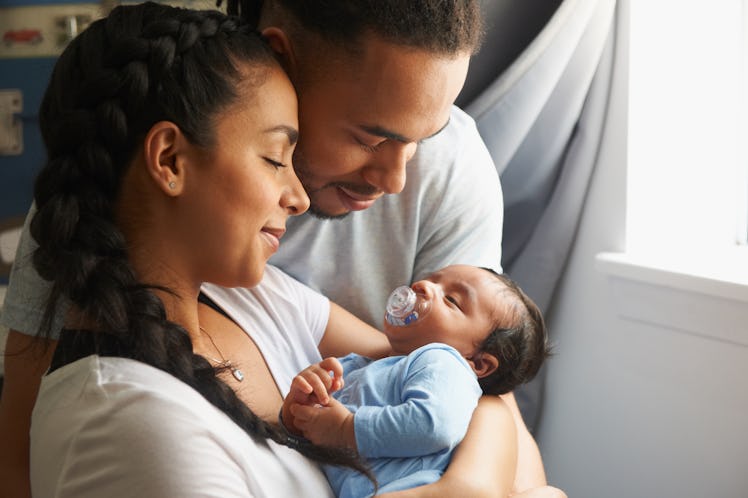 A new mom and dad cradle their newborn baby in their arms as the baby sucks on a pacifier.