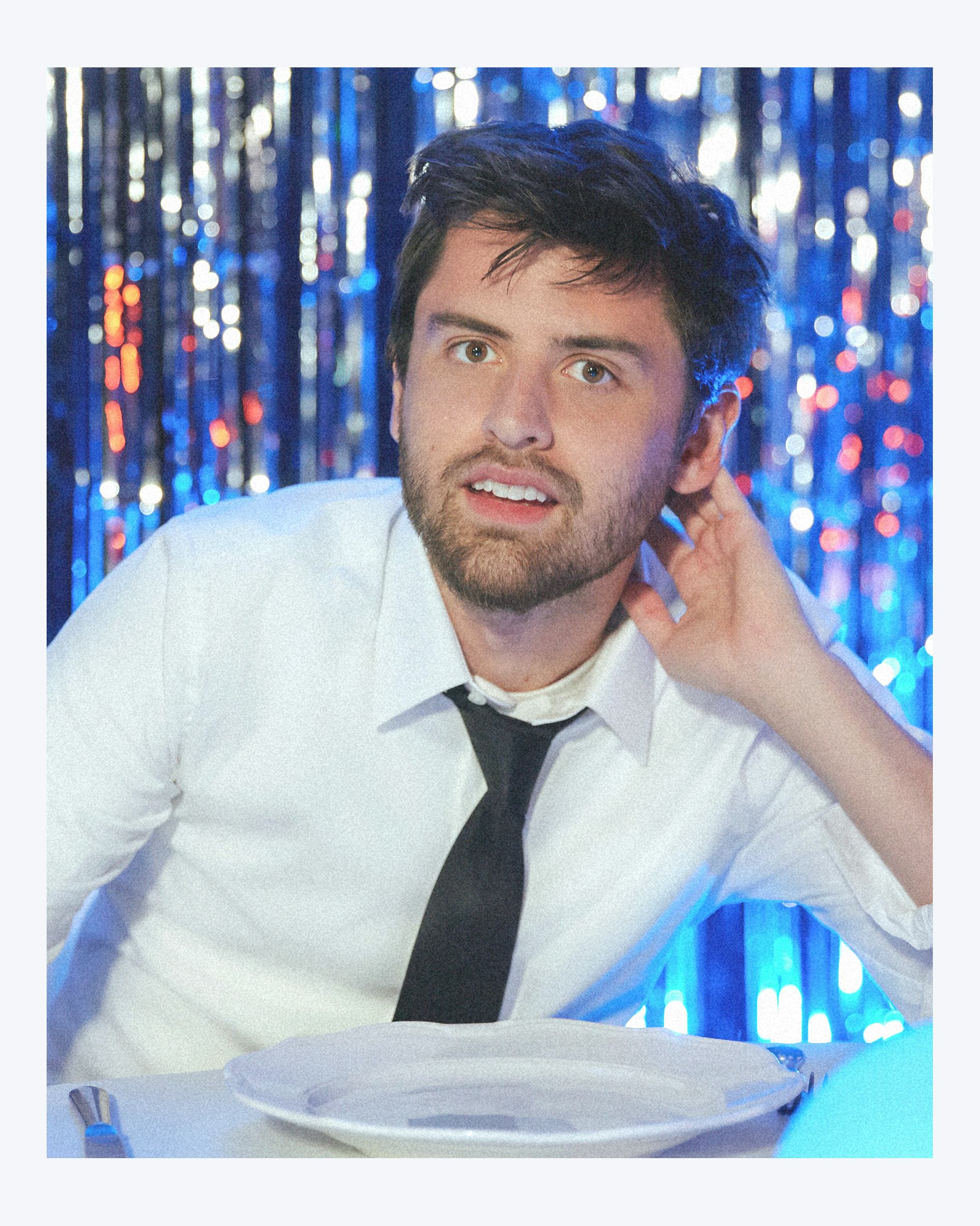 A portrait of Cooper Raiff in front of a wall of blue shiny tinsel.