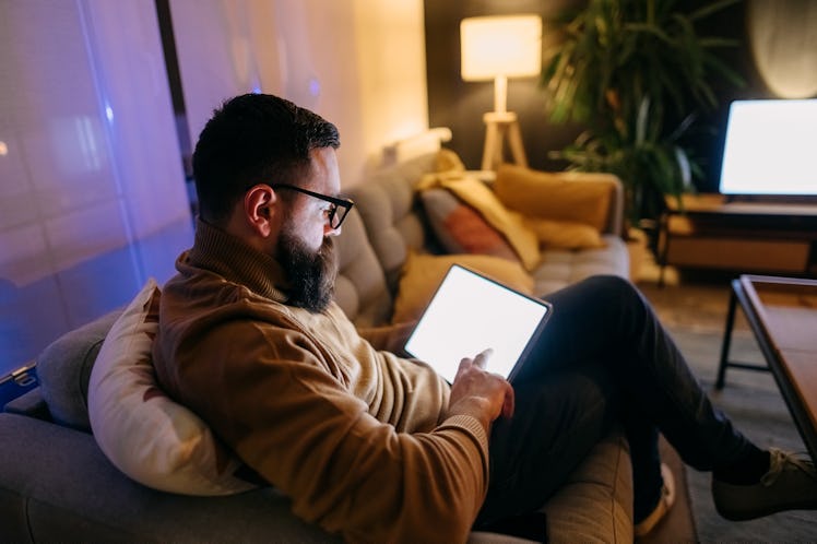 A man in a yellow sweater sitting on a couch in his living room scrolling on his tablet