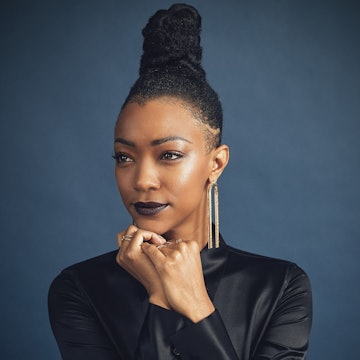 Sonequa Martin-Green wearing a black satin shirt and gold jewelry with her hair tied up and hands un...