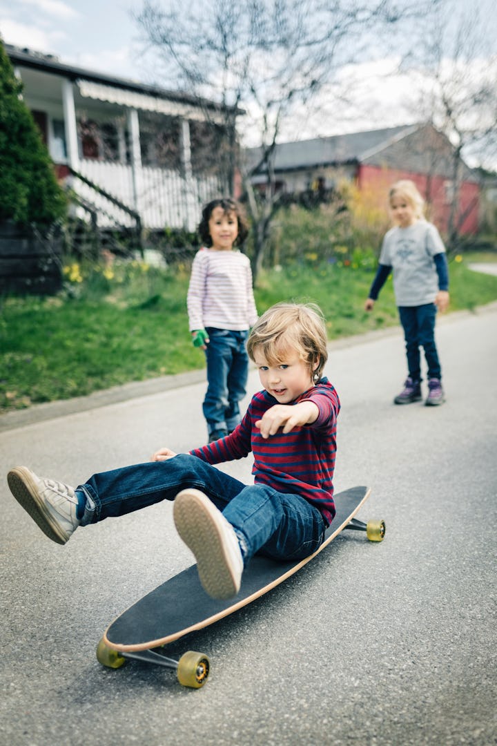 A young boy sits on a skateboard while his siblings look on. 