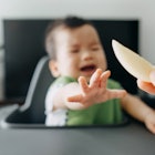 A crying baby in a high chair reaching for an apple slice that their parent is holding in a pincer g...