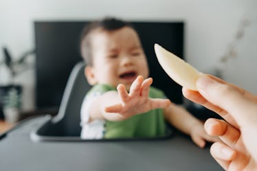 A crying baby in a high chair reaching for an apple slice that their parent is holding in a pincer g...