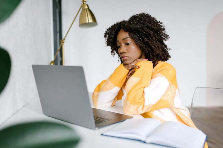 An unhappy woman sitting by the desk and looking at her laptop while trying to deal with a toxic wor...