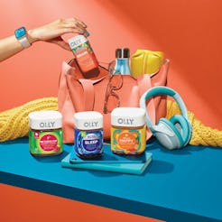 A hand putting Olly’s vitamin gummies in the bag standing next to boxes of Olly vitamins, headphones…