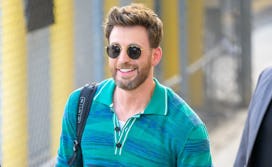 Chris Evans in sunglasses with a greying beard in a striped blue dress shirt smiling on a sunny day