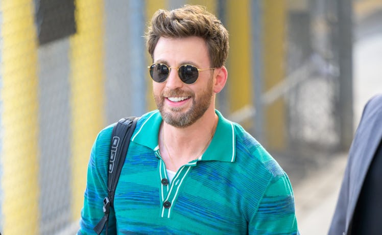 Chris Evans in sunglasses with a greying beard in a striped blue dress shirt smiling on a sunny day