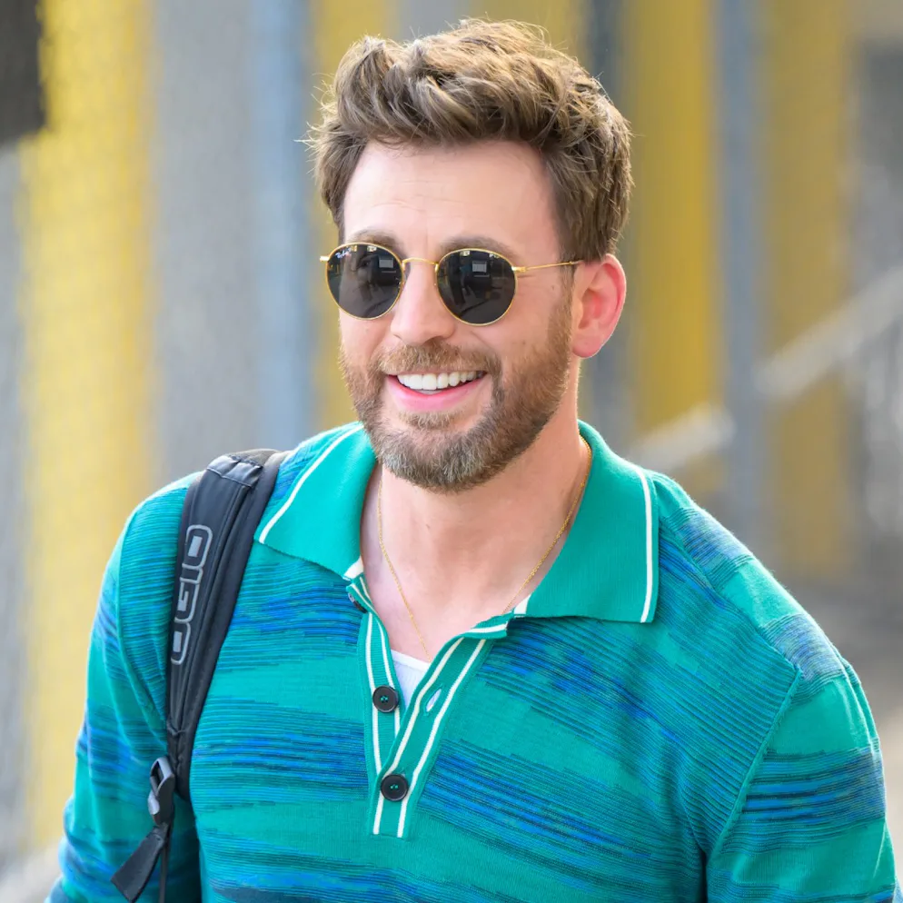 Chris Evans in sunglasses with a greying beard in a striped blue dress shirt smiling on a sunny day