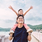A girl with her arms raised, sitting on her dad's shoulders and smiling as he stands on a bridge.