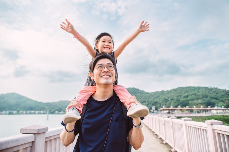 A girl with her arms raised, sitting on her dad's shoulders and smiling as he stands on a bridge.
