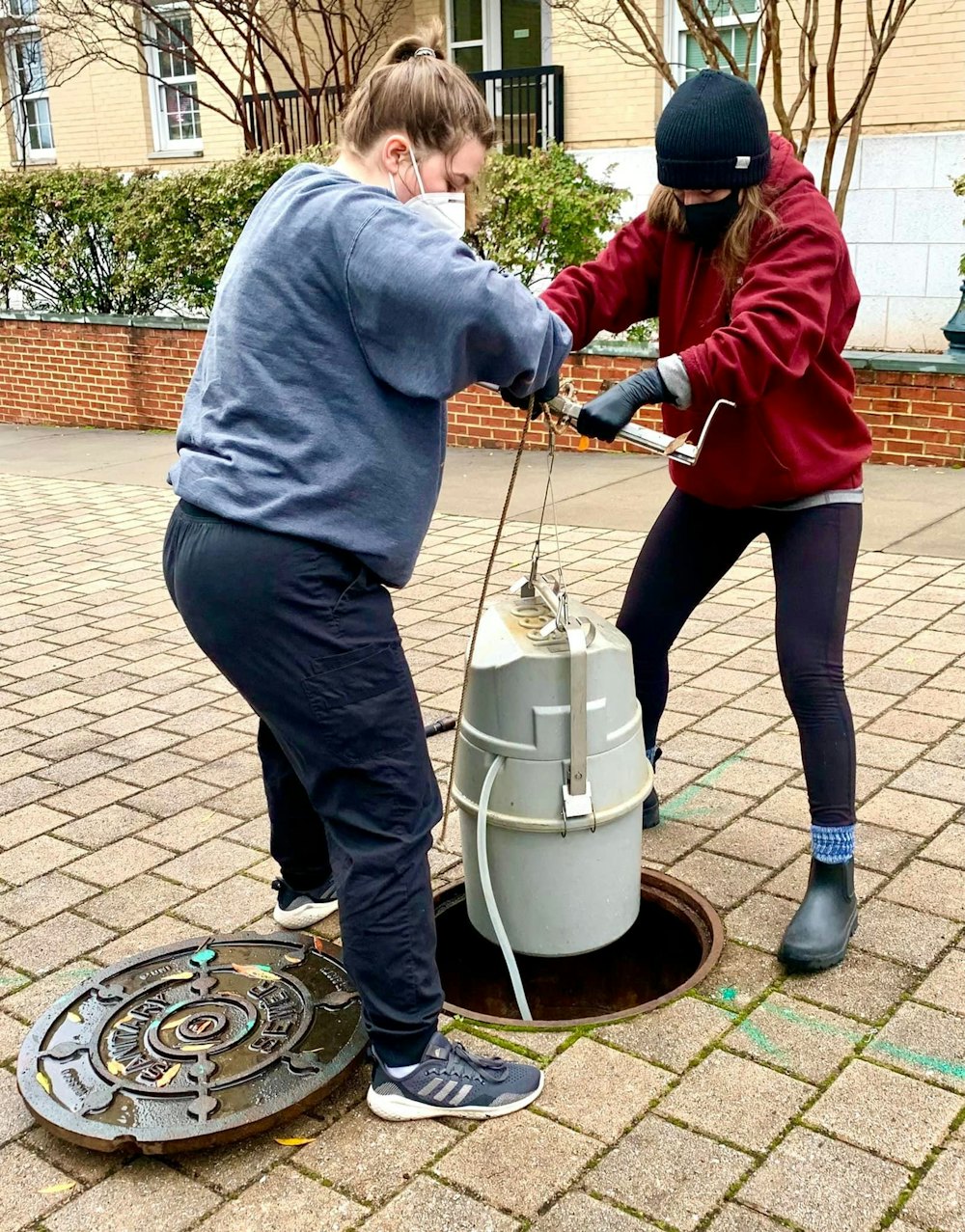 Two students wearing masks lower a Shop-Vac sized container into an open manhole.