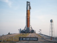 The Space Launch System rocket sits atop a small hill in front of a banner that reads "WE ARE GOING"