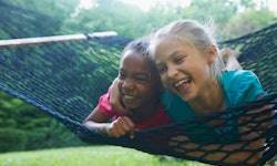 A Black girl and white girl swing on a hammock on their stomachs, laughing together.