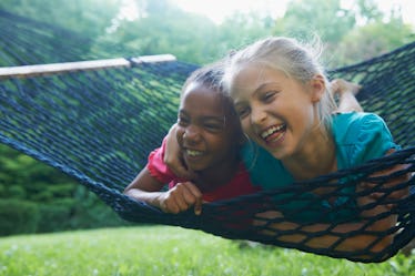 A Black girl and white girl swing on a hammock on their stomachs, laughing together.