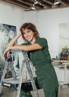 The artist Emma Webster standing on a ladder in her studio, smiling at the camera