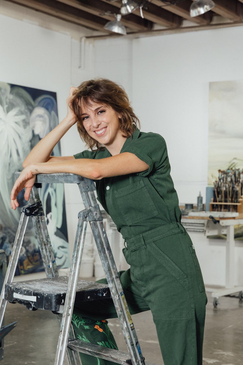 The artist Emma Webster standing on a ladder in her studio, smiling at the camera