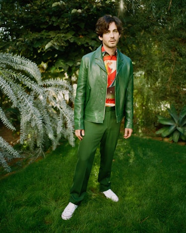 Logan wearing green jacket and green pants, standing in a woodsy area.