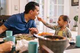 A dad opens his mouth for his daughter to feed him a bite of food at the dinner table.