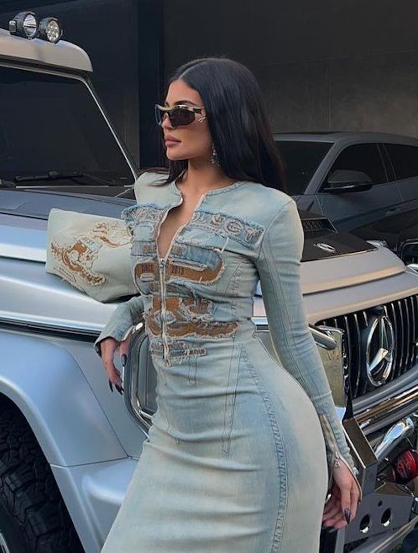 A Closeup of Kylie Jenner posing in front of a silver Mercedes-Benz G-Class in a full denim dress.