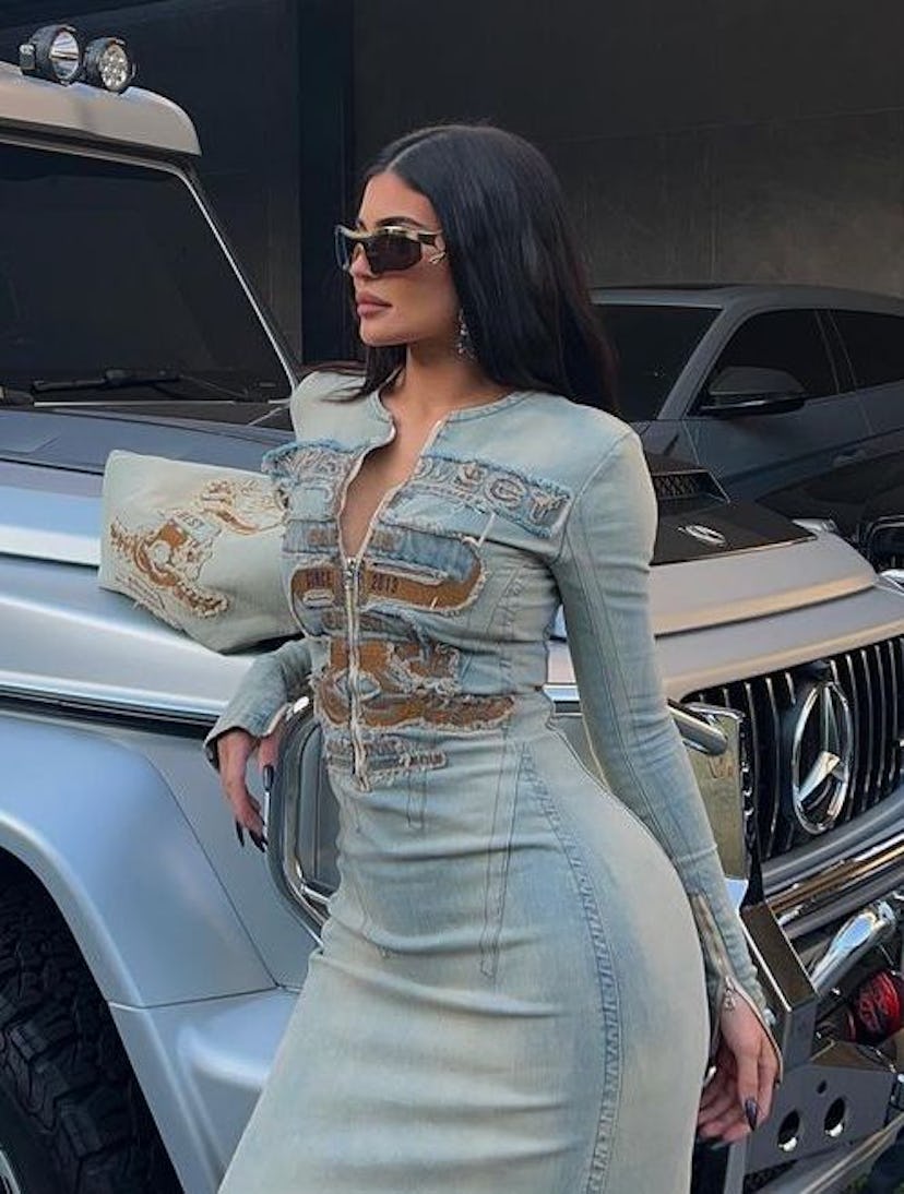 A Closeup of Kylie Jenner posing in front of a silver Mercedes-Benz G-Class in a full denim dress.