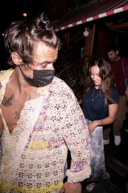 Harry Styles in a loose embroidered shirt with Olivia Wilde walking behind him and smiling 