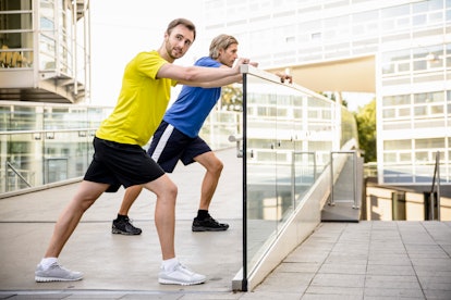 Two men stretch their calves with their front leg bent and back leg in a lunge and straight, heel on...