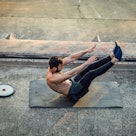 A man does a v sit-up outdoors on cement on a yoga mat.