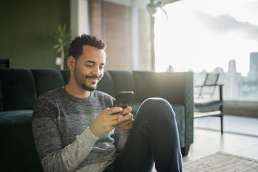 Man sitting on floor with knees up using his phone sending a thoughtful text to his stressed out par...