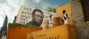 Two Wakandan drummers stand next to a mural of T'Challa (Chadwick Boseman) in Black Panther: Wakanda...
