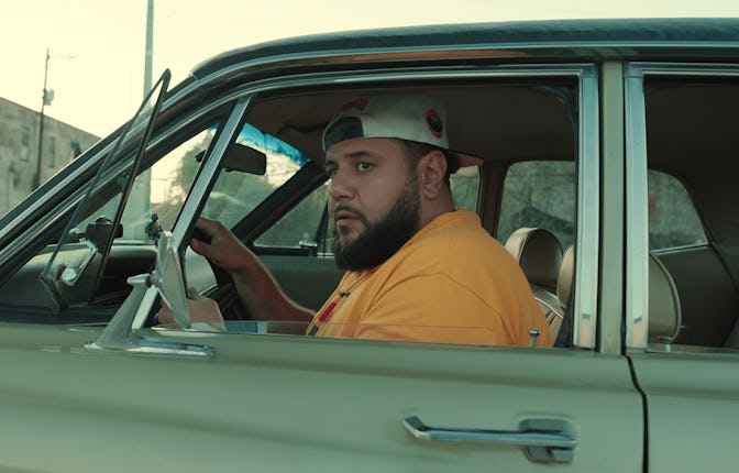 Mo Amer sitting in a car, with rolled down windows, hands on the steering wheel, looking through the...