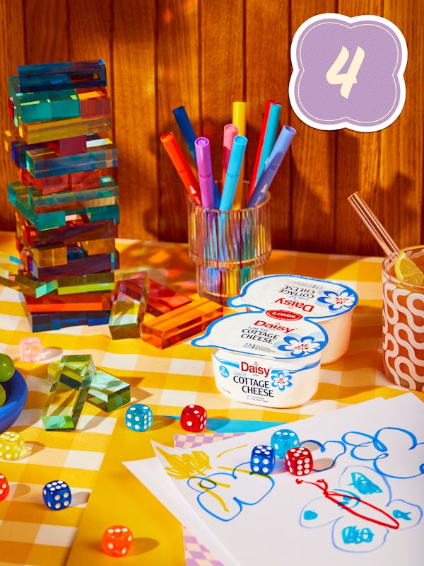 Two low fat Daisy Cottage Cheeses on a table along with children's doodles on papers, dice and color...