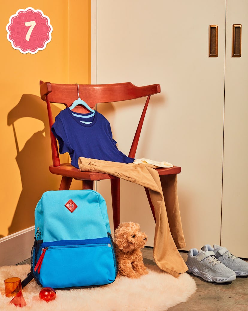 A blue shirt and beige pants hanging on a wooden chair, grey sneakers on the floor and a blue backpa...