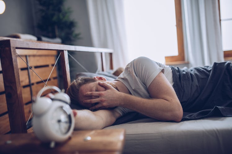 A man with a hangover in bed, hand covering his face, as his alarm clock goes off.