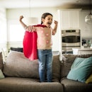 A child in a cape flexing their biceps and smiling while standing on a couch at home.