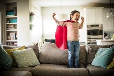 A child in a cape flexing their biceps and smiling while standing on a couch at home.
