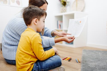 A child getting evaluated for autism sits on a floor with a psychologist who is holding up a piece o...
