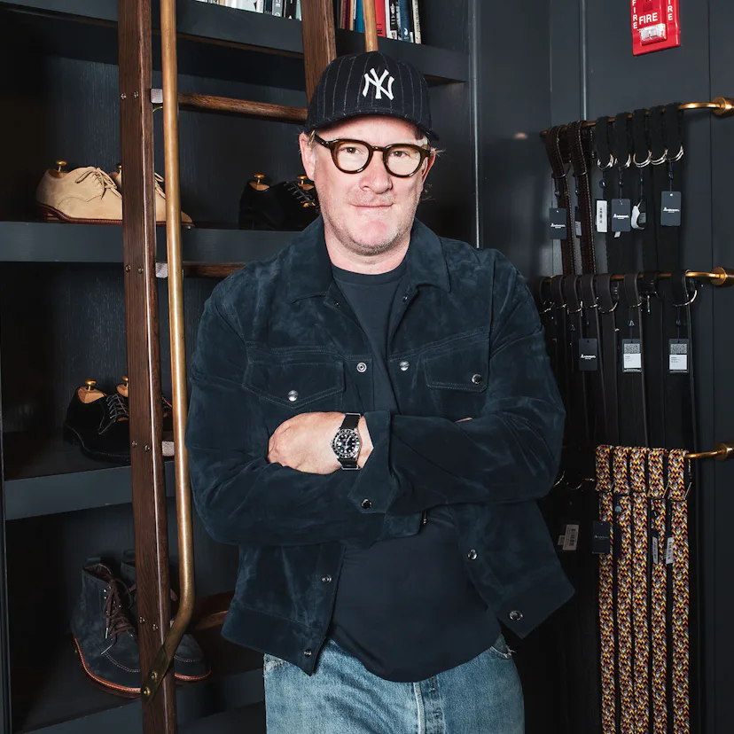 Todd Snyder wearing a new york yankees hat standing next to a rack full of belts and shelves of shoe...