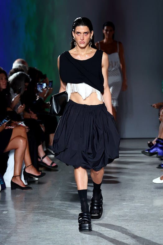 A model wears a voluminous black skirt paired with an asymmetric black crop top, accessorized with w...
