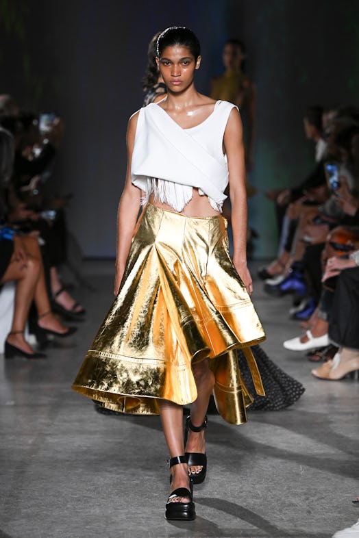 A model wears a fringed white crop top with asymmetric draping and a voluminous metallic skirt by Pr...