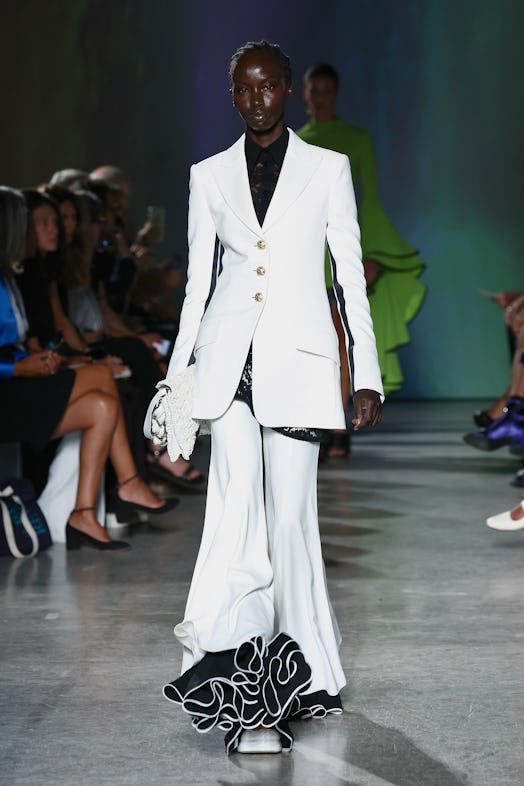 A model wears a white suit, pairing a long gold-buttoned blazer and bell-bottom trousers with large,...