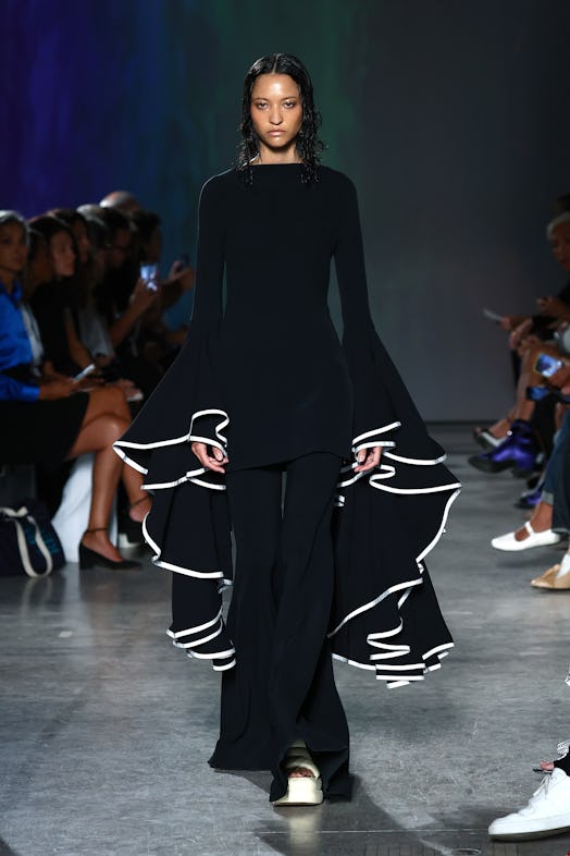 A model wears a black tunic featuring white-trimmed oversized bell sleeves paired with black trouser...