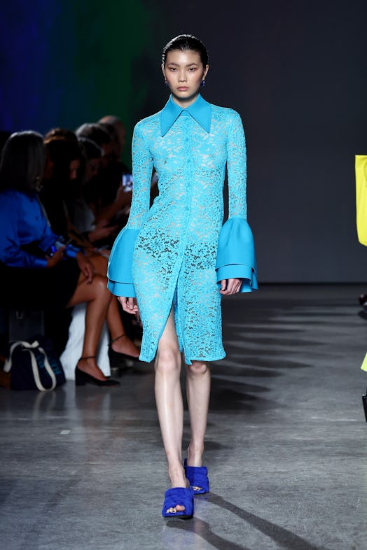 A model wears a bright see-through lace dress paired with dark, royal blue heeled slip-on sandals by...